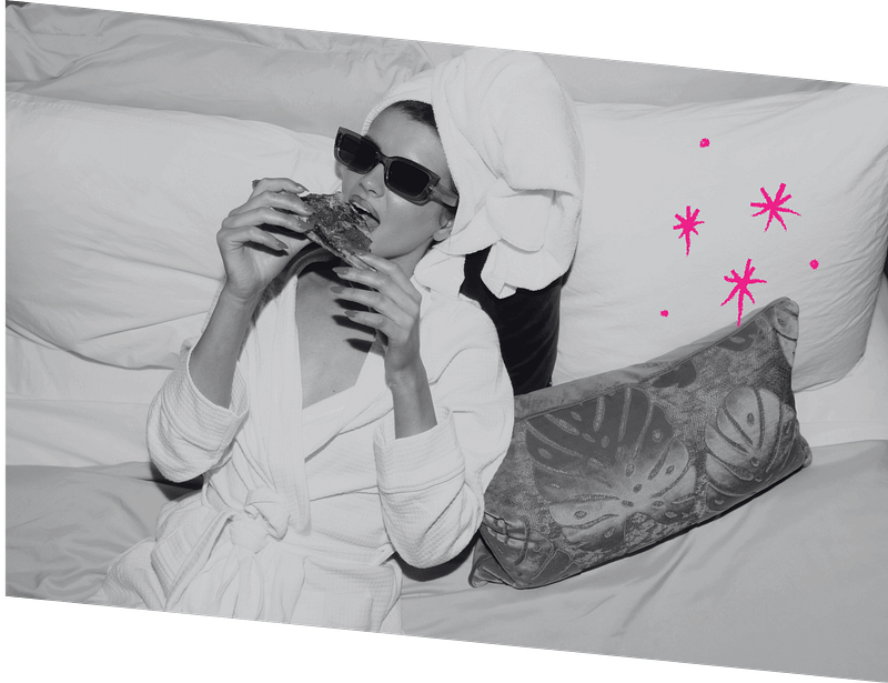Person in a bathrobe and sunglasses, with a towel on their head, relaxes on a couch while eating pizza. Pink star doodles are on the right.