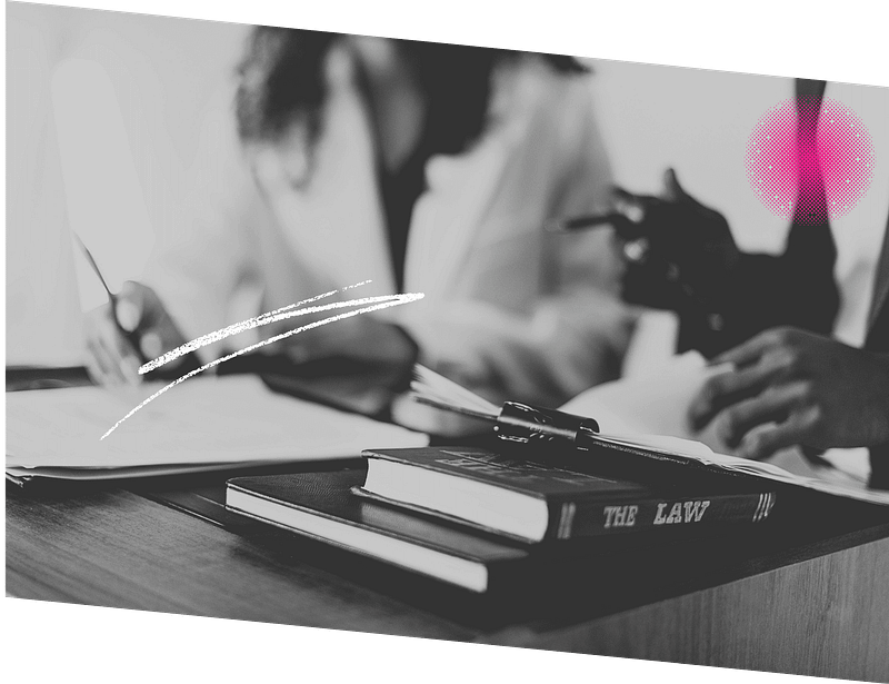 Two people seated at a table, with law books and documents. They appear to be discussing or reviewing paperwork. The image is in black and white with a pink accent.